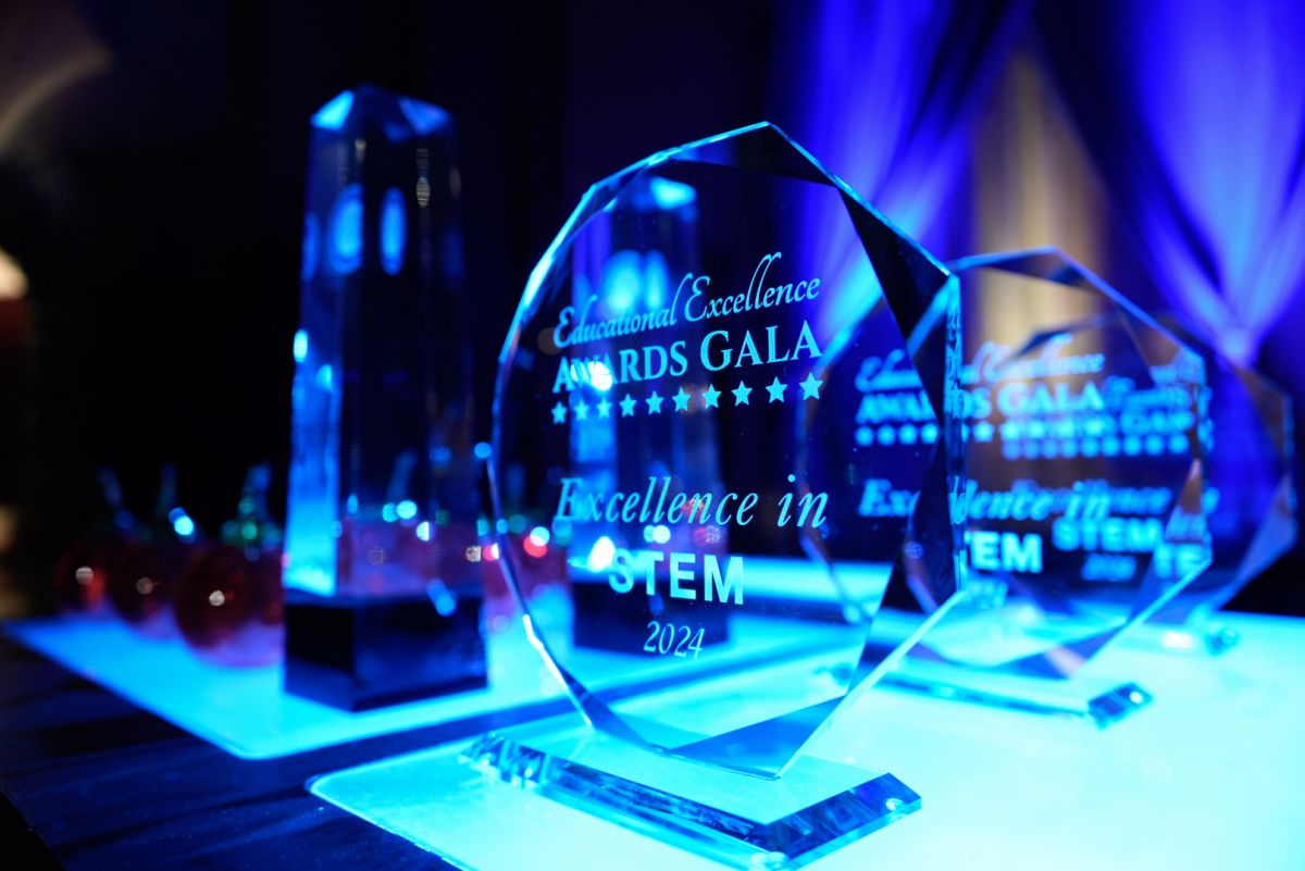 On a table lit with blue light are several trophies for the Educational Excellence Awards. The awards are transparent and have white text and the gala logo.