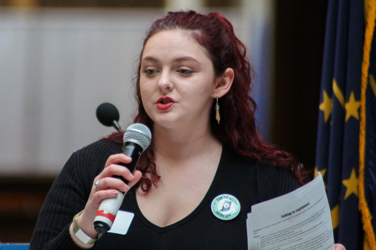 Eliza Brader speaks into a microphone at a rally in the Indiana Statehouse. She wears a black shirt and has her hair pulled back. Behind her is an Indiana state flag.