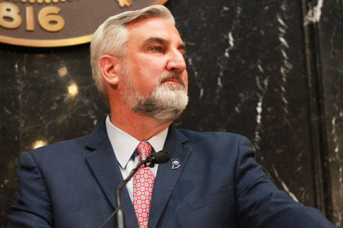 Eric Holcomb stands in front of a microphone in the Indiana House. Holcomb is a White man with white and gray hair and beard. He is wearing a blue suit.