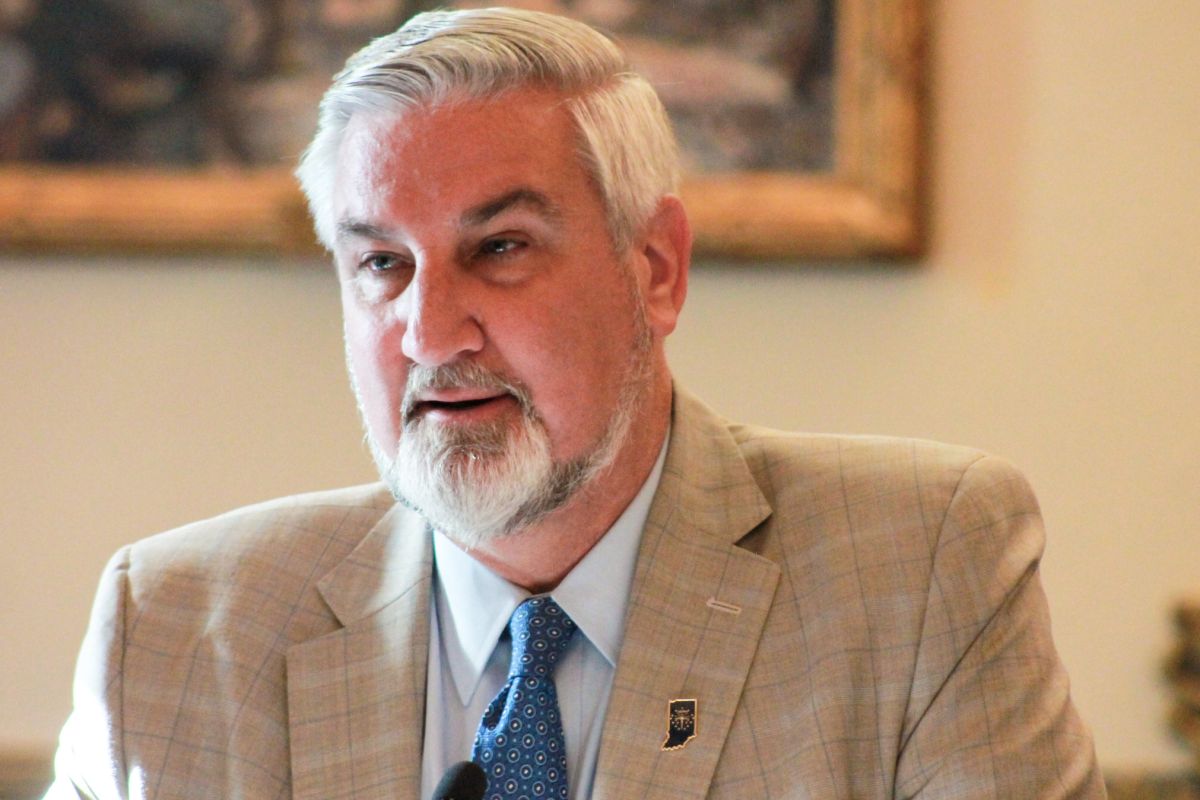 Eric Holcomb is a White man with white hair and beard. He is wearing a tan suit and blue tie.