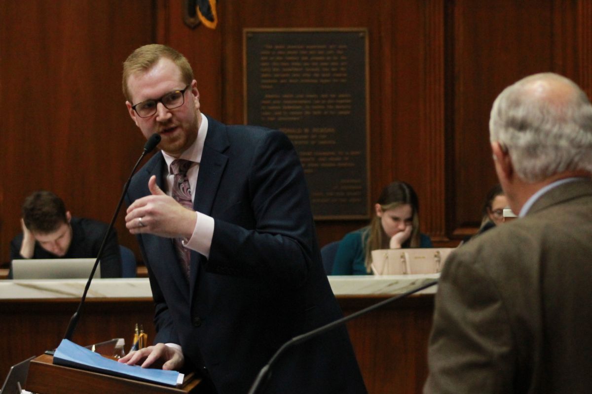 Reprsenative Ethan Manning wearing a navy blue suit with a white shirt and a paisley patterned tie speaking into a microphone while debating another lawmaker while on the House floor.