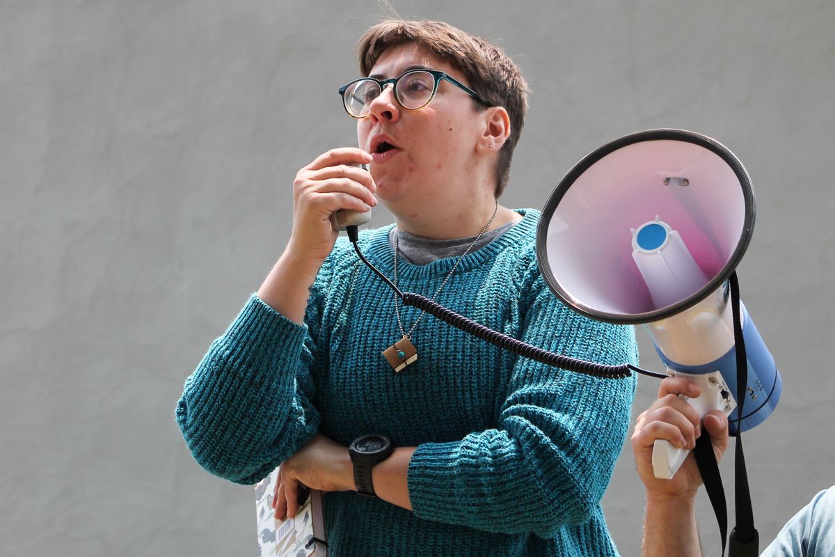 Evie Phelps speaks into a transmitter for megaphone. The megaphone held by another person off-camera.