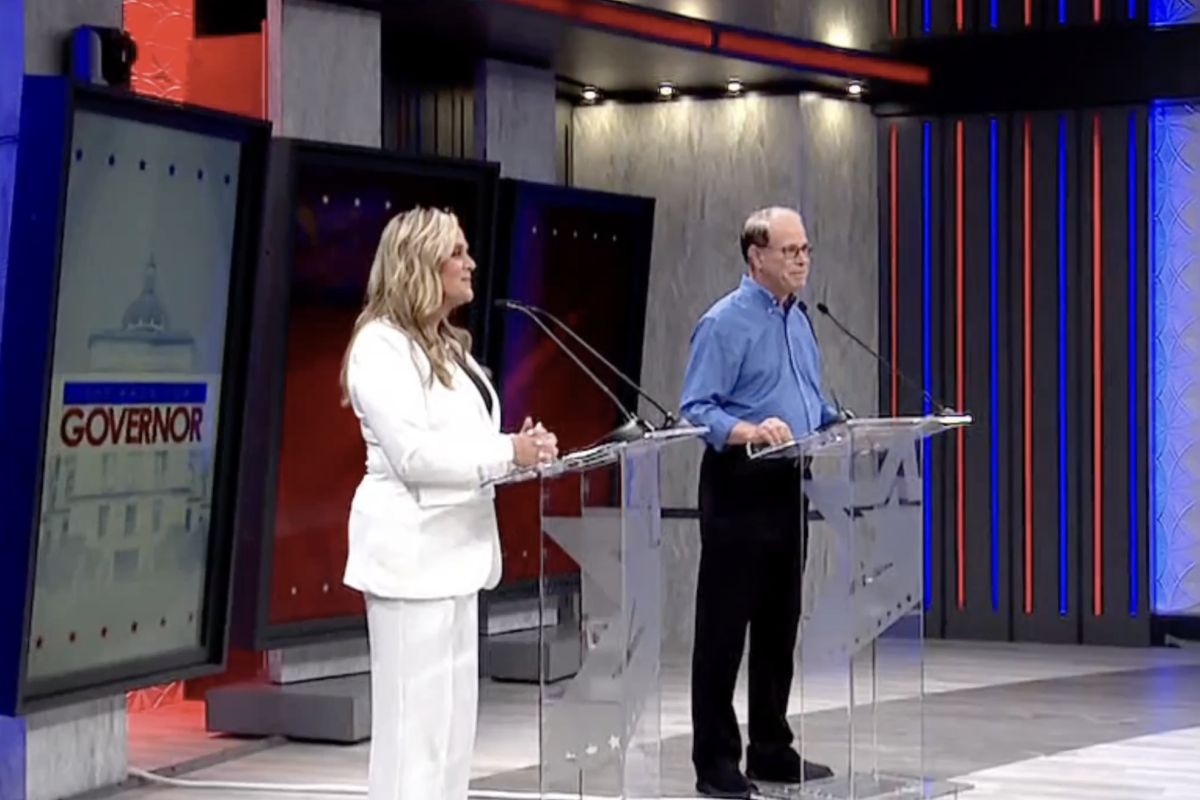 A screenshot of a televised gubernatorial debate. Jennifer McCormick is a White woman with blonde hair. She is wearing a white pantsuit. Mike Braun is a White man with dark, graying hair. He is wearing glasses, a blue shirt and black pants.