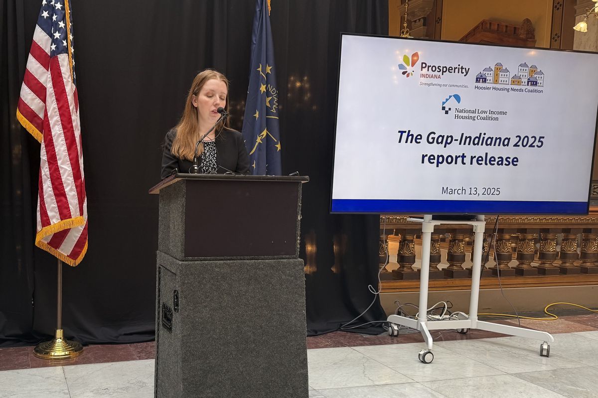 There is an American flag in front of a black curtain. There is a white woman with blonde hair speaking at a podium with a nearby screen that reads "The Gap-Indiana 2025 report release". 