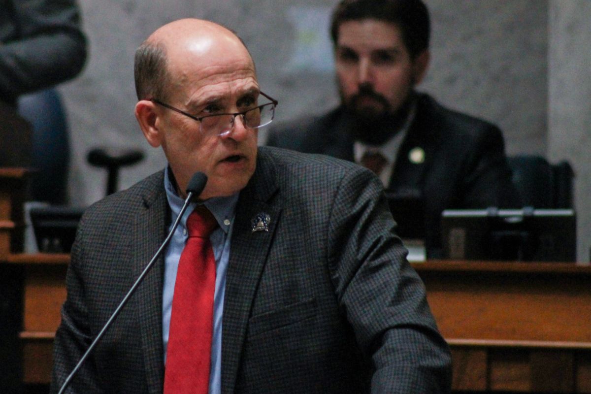 Gary Byrne speaks into a microphone at a lectern in the Senate. Byrne is a White man, mostly bald. He is wearing reading glasses and a dark suit with a blue shirt and a red tie.