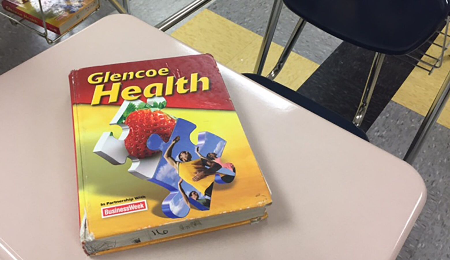 A red and yellow textbook titled "Glencoe Health" with puzzle pieces on the front is displayed on a desk in a school classroom.