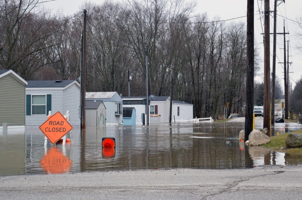 A row of homes sits in flooded waters. A sign in the foreground reads ROAD CLOSED.