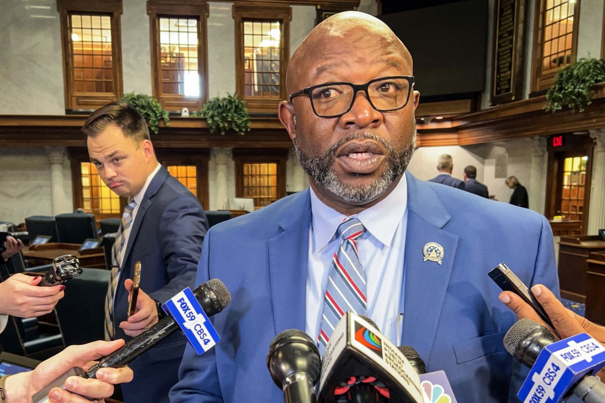 Greg Taylor speaks in front of a bank of microphones. Taylor is a Black man, bald with a dark, graying beard. He is wearing glasses and a suit and tie.