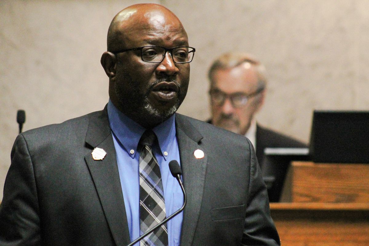 Greg Taylor is a Black man, bald, with a white goatee. Taylor is wearing glasses and a suit and tie.