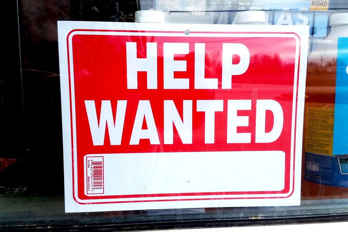 A red sign with a white border reads "HELP WANTED" in capital bold white letters inside of a window. 