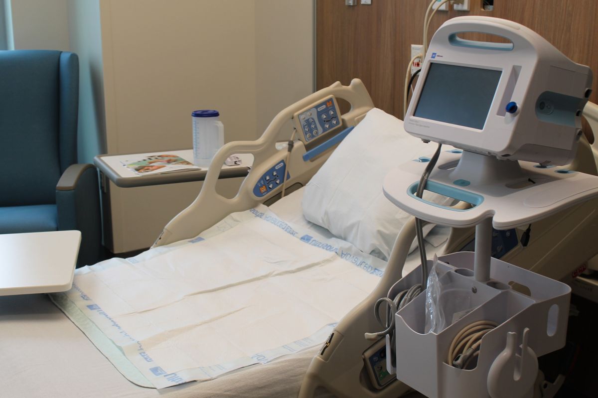 A medical monitor sits next to a hospital bed with a pillow and pad on it in a patient room.