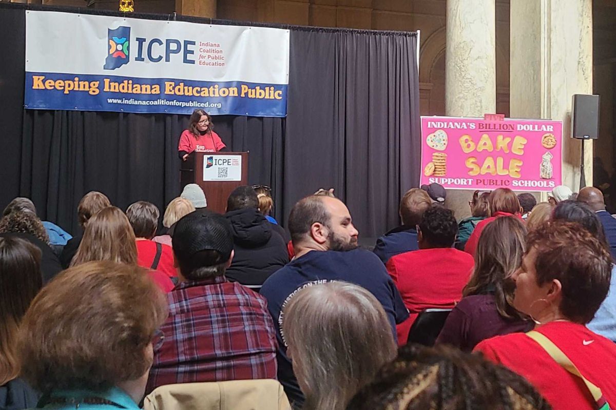 Educators and public education supporters gather in seats in front of a person speaking at a podium. A hot pink sign reads "bake sale" and a banner behind the speaker says "Keeping Indiana Education Public." 