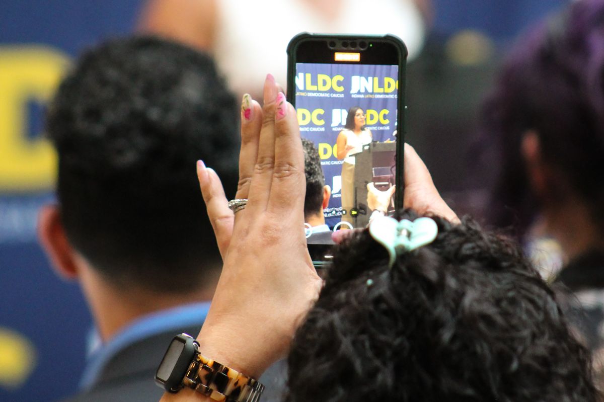 The Indiana Latino Democratic Caucus vice president speaks at a podium as a member of the audience records her with their phone. The audience member is facing away from the camera, but they have dark curly hair in a claw clip and a tortoiseshell watc