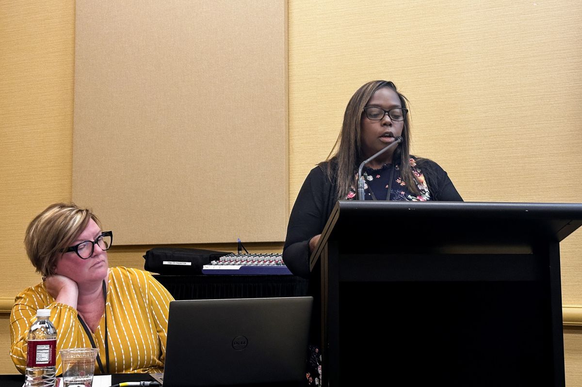 Ashley Coleman speaking into a microphone at a podium. Kristen Moore is sitting down next to her as part of a panel. 