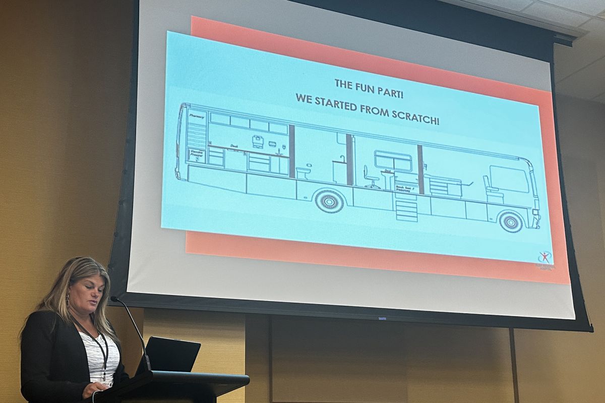 Joyce Gies speaking in front of a screen with a projection of a presentation of Rush Memorial Hospital's mobile clinic.