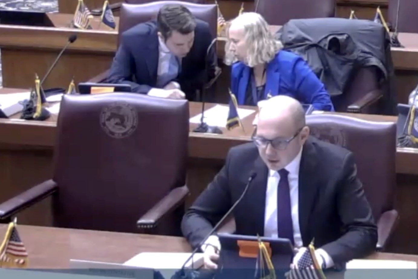 Jake Teshka sits on a red chair at a desk framed by small Indiana and United States flags. He is a White man, bald, wearing a suit and glasses. Two people whisper at the desk behind him. 