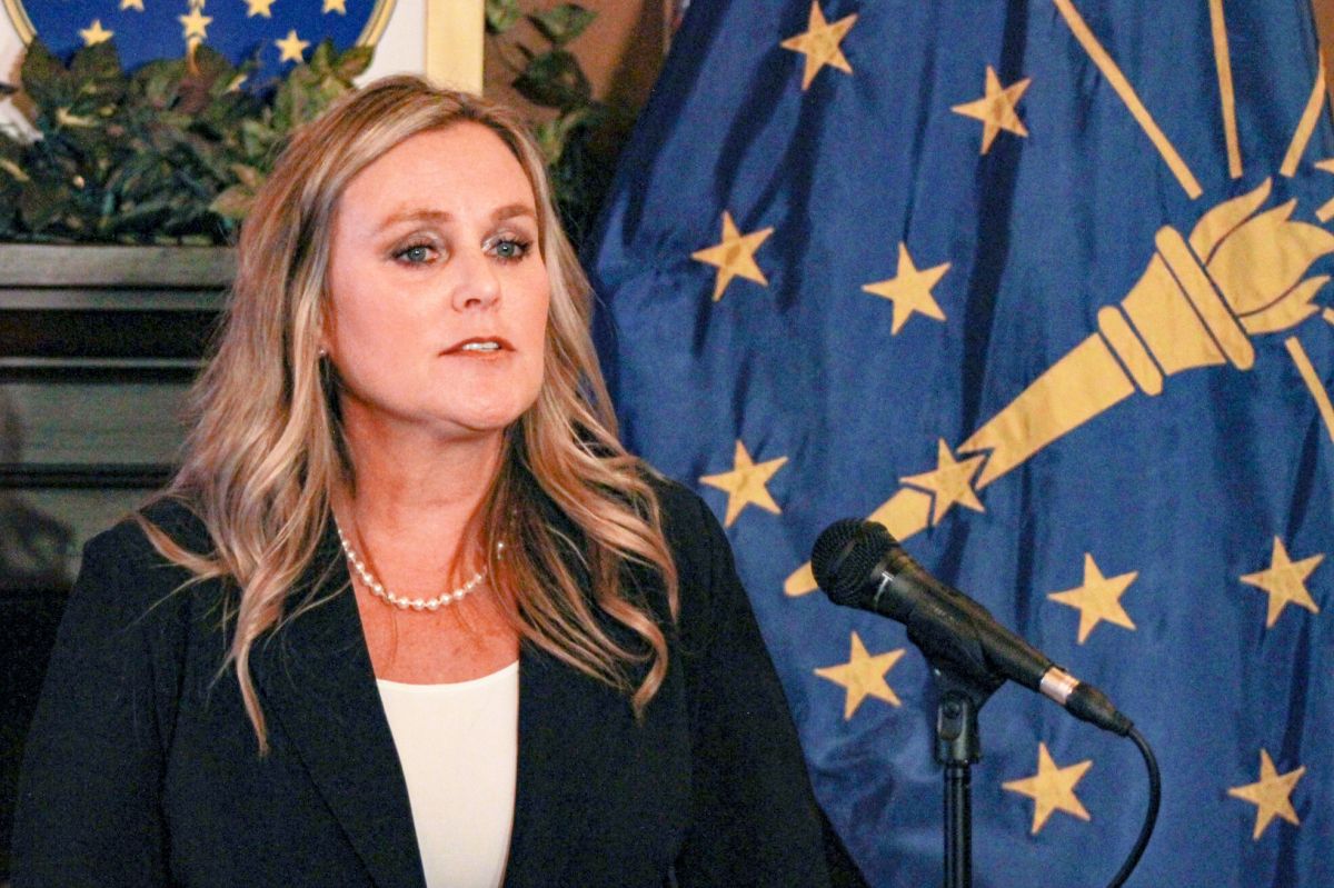 Jennifer McCormick speaks into a microphone with the Indiana flag draped behind her. McCormick is a White woman with blonde hair. She is wearing a black jacket over a white top.