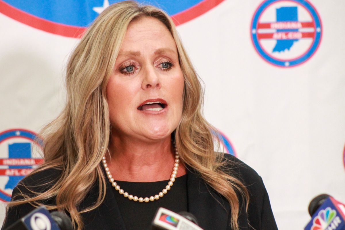 Jennifer McCormick speaks into a bank of microphones during a press conference. McCormick is a White woman with blonde hair. She is wearing a black jacket over a black top.