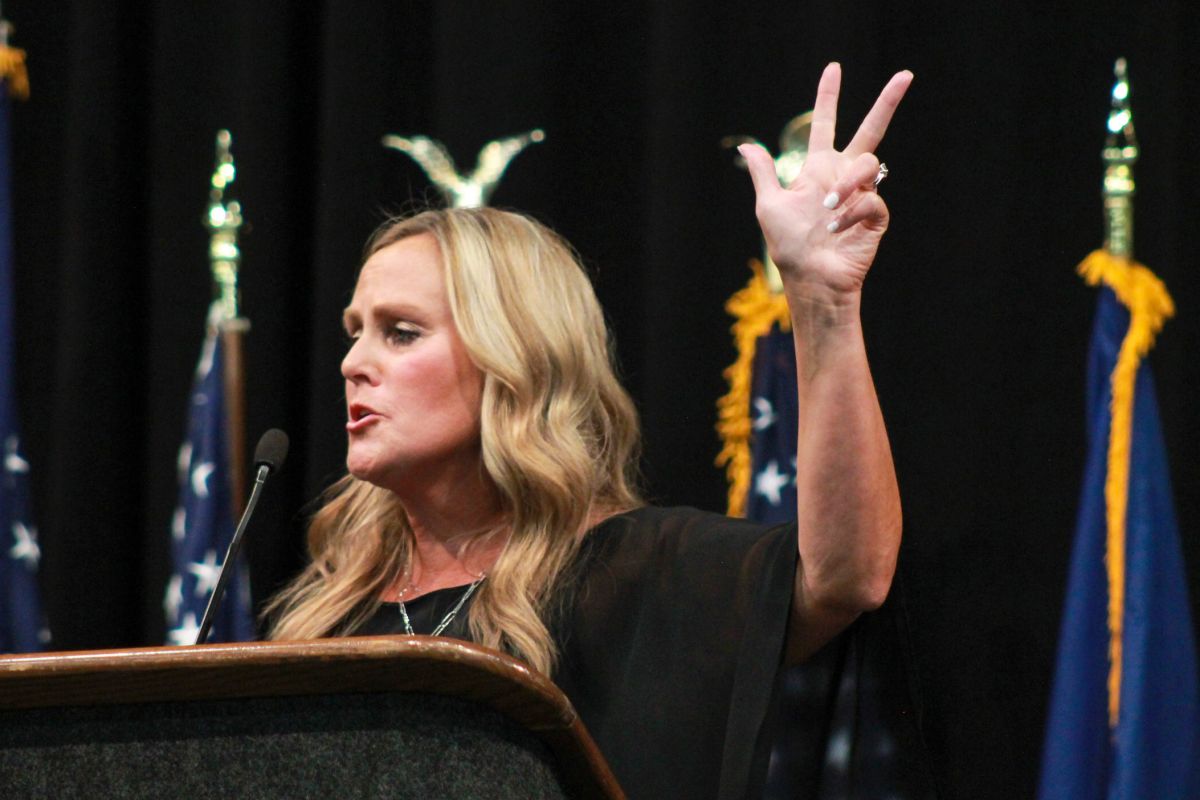 Jennifer McCormick holds her hand in the air, gesturing as she speaks at a lectern. There are a row of flags behind her. McCormick is a White woman with blonde hair. She is wearing a black top.