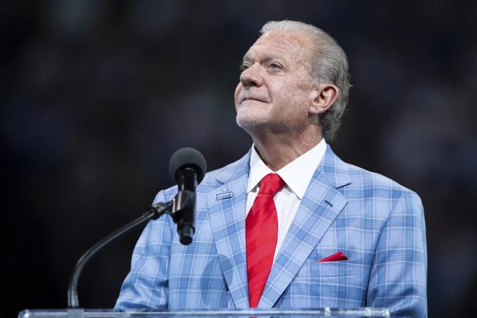 Jim Irsay, a white man with gray hair, speaking at a podium. He's wearing a light blue plaid suit, white shirt and red tie.