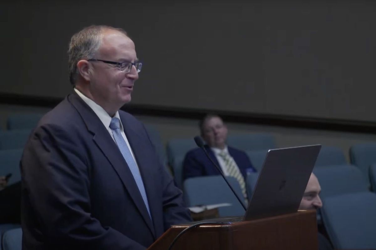 John Keller stands at a podium and speaks into a microphone. Keller is a White man and he is wearing a dark suit with a light blue necktie.