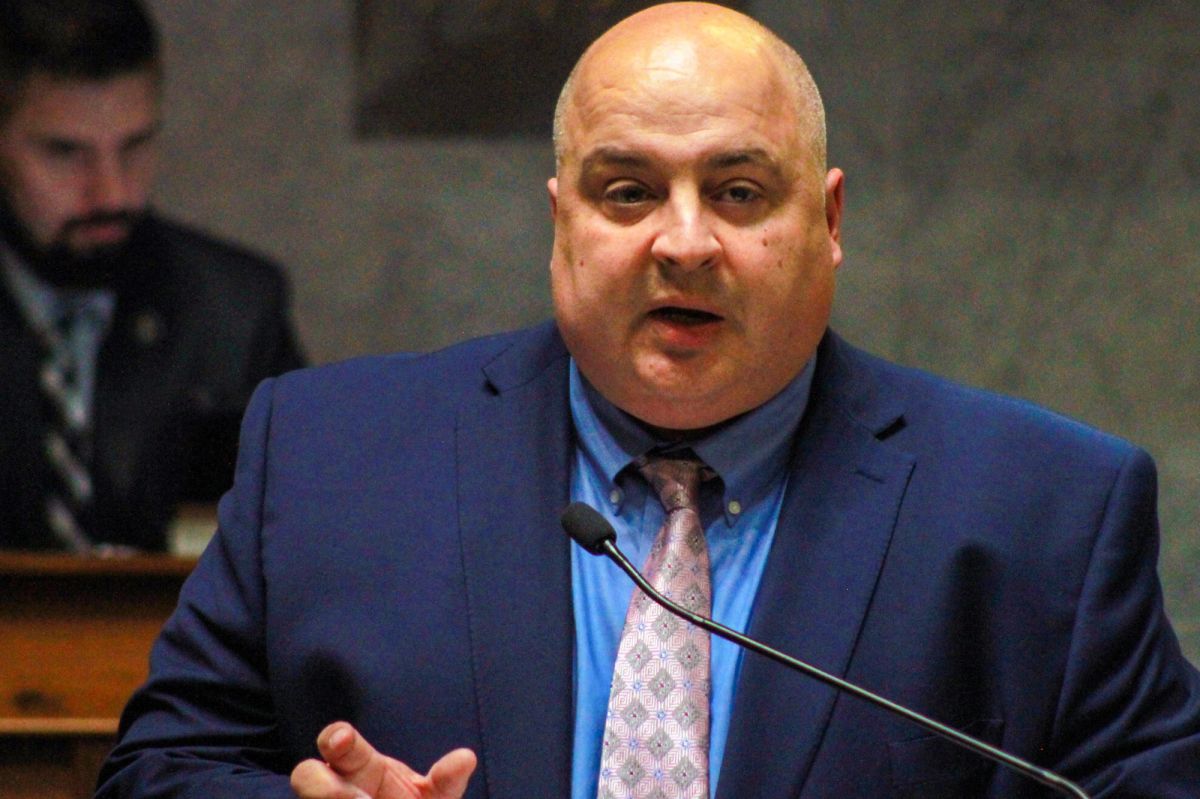 Senator Jon Ford testifies on the floor of the Indiana Senate. He is a White man, bald wearing an navy suit. He stands in front of a microphone.