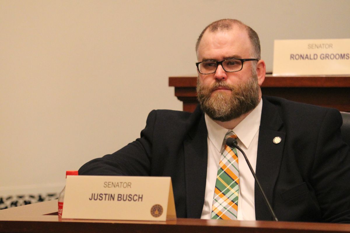 Senator Justin Busch in a suit and tie, sitting in a committee room. Busch is a White man with a graying brown beard and short, graying brown hair.