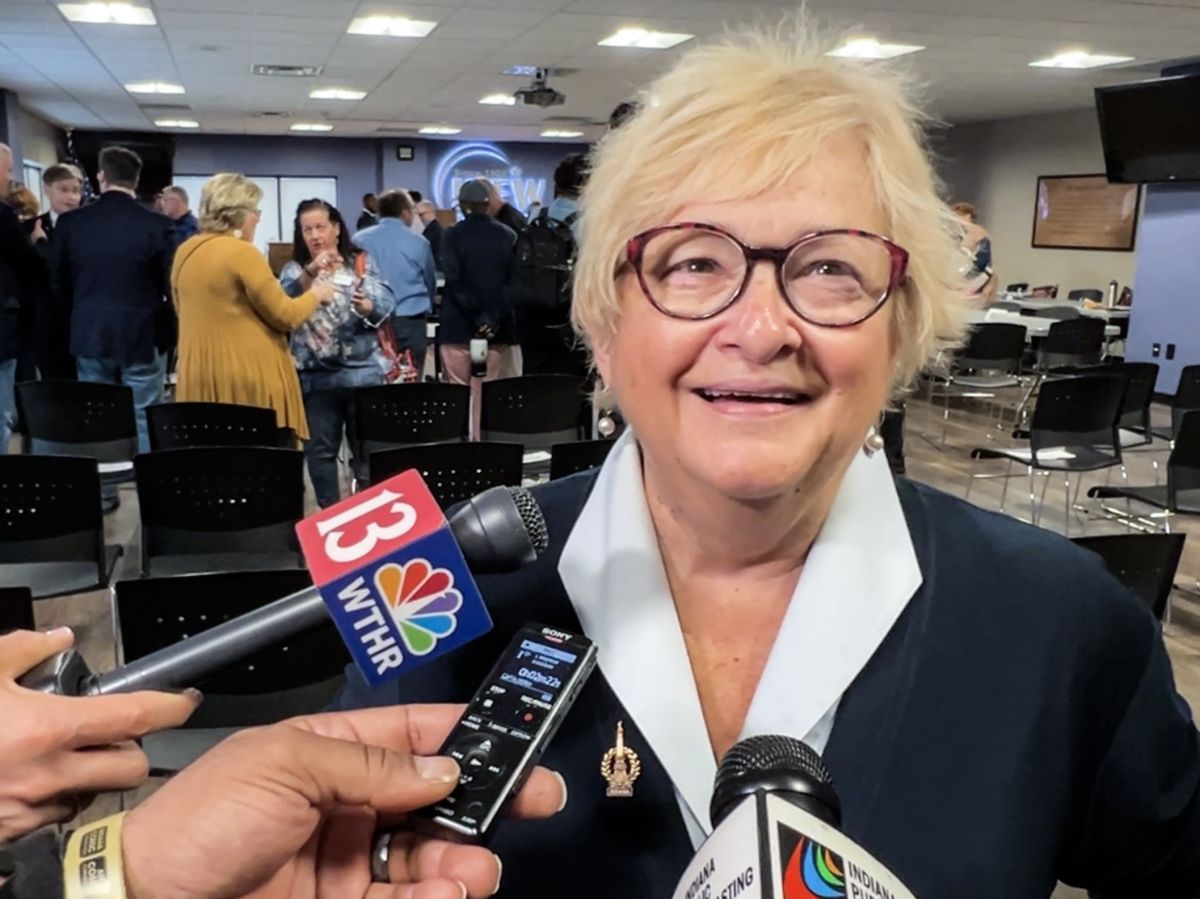  karen talian is white with blonde hair. She is wearing a black suit talking into a microphone.