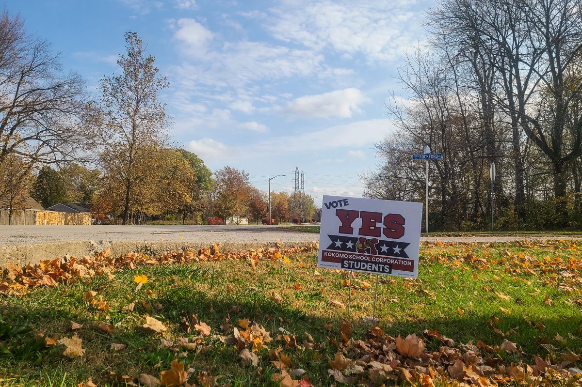 A yard sign supporting Kokomo's school referendum is displayed in the grass of a lawn with scattered foliage. Behind it is an intersection of a neighborhood.