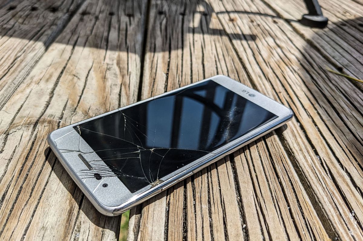 A phone with a cracked screen rests on a wooden deck. The photographer would like you to know this is the first screen she's cracked since 2006.