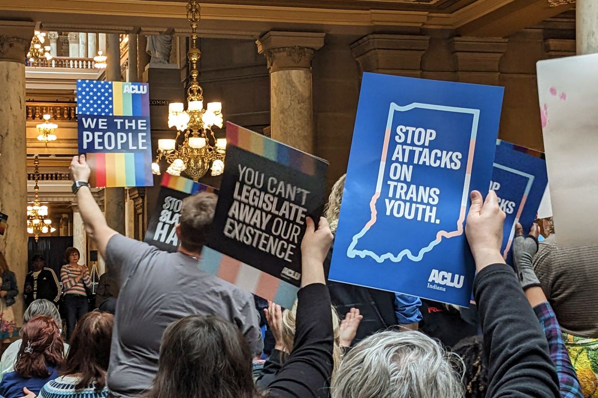  A crowd holds signs at Indiana's 2023 LGBTQ+ Statehouse Day. The sign in focus reads 'Stop attacks on trans youth.' The text is in the center of an outline of Indiana in the colors of a trans pride flag. 