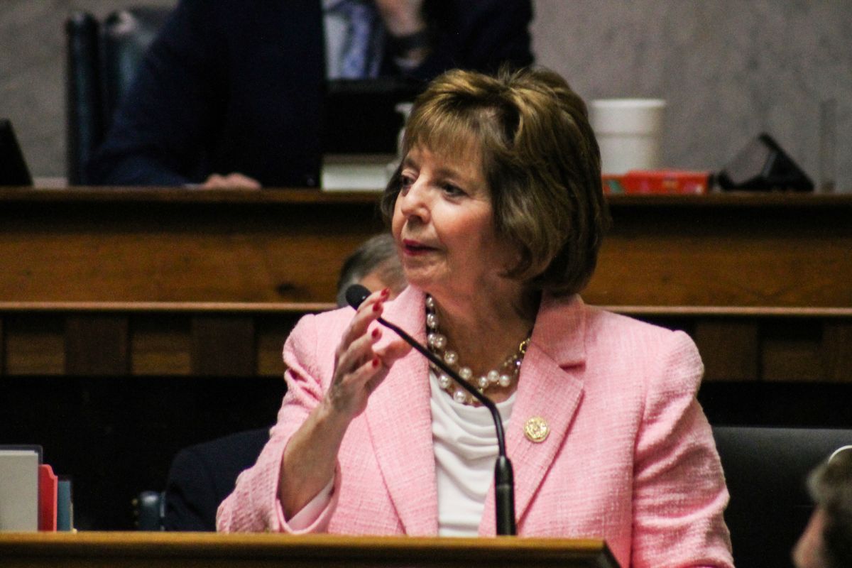 A woman in a light pink blazer wearing a gold pin and pearls turns her head and lifts her right hand as she speaks into a microphone in the Indiana Senate Chamber.