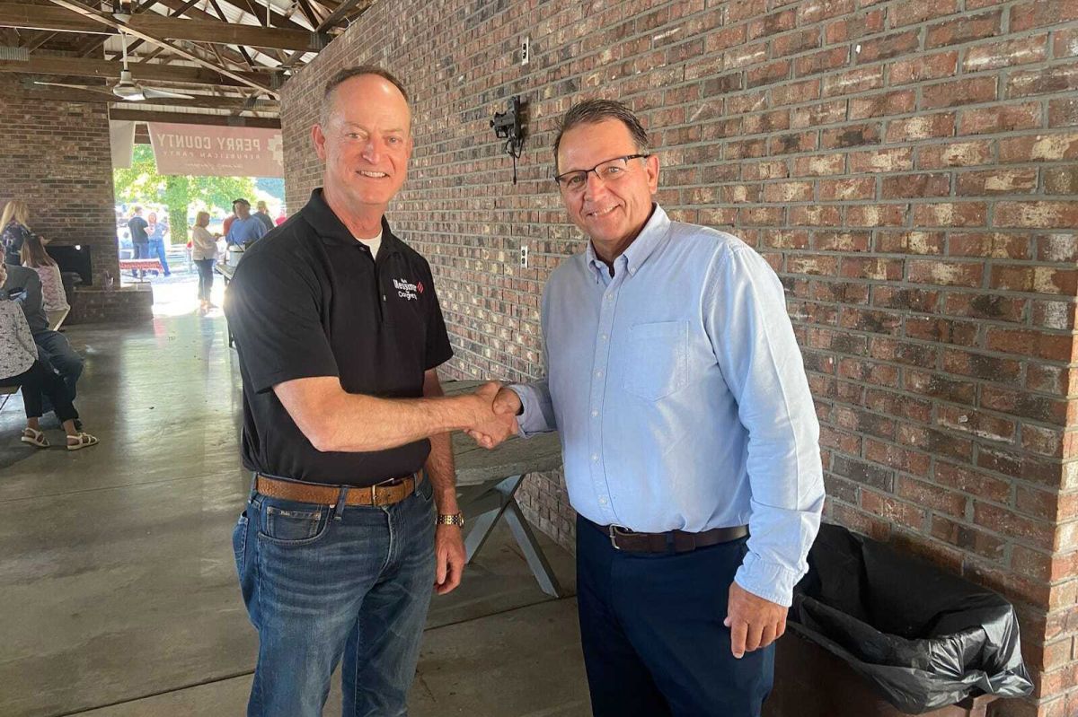 Mark Messmer shakes hands with Derek Schmitt. Messmer and Schmitt are both White men with dark hair. Messmer is wearing a polo shirt and jeans. Schmitt is wearing glasses, a button-down shirt and jeans.