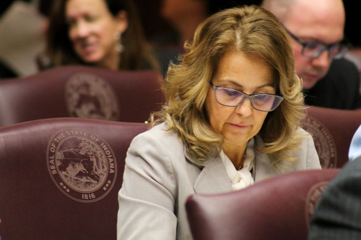 Representative Michelle Davis sits at her desk on the floor of the Indiana House. She is a White woman with glasses, wearing a grey suit.