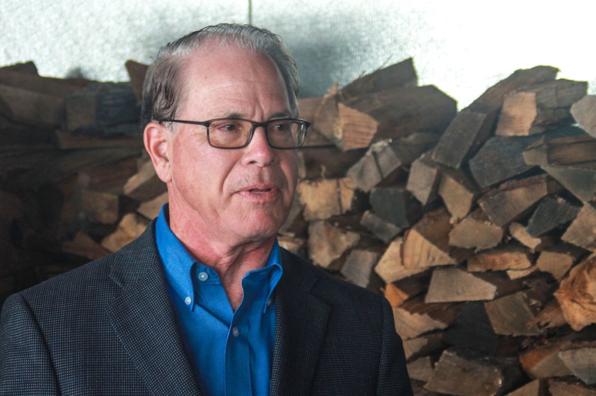 Mike Braun stands in front of a pile of firewood. Braun is a White man with dark, graying hair. He is wearing glasses and a gray sport coat over a blue shirt.