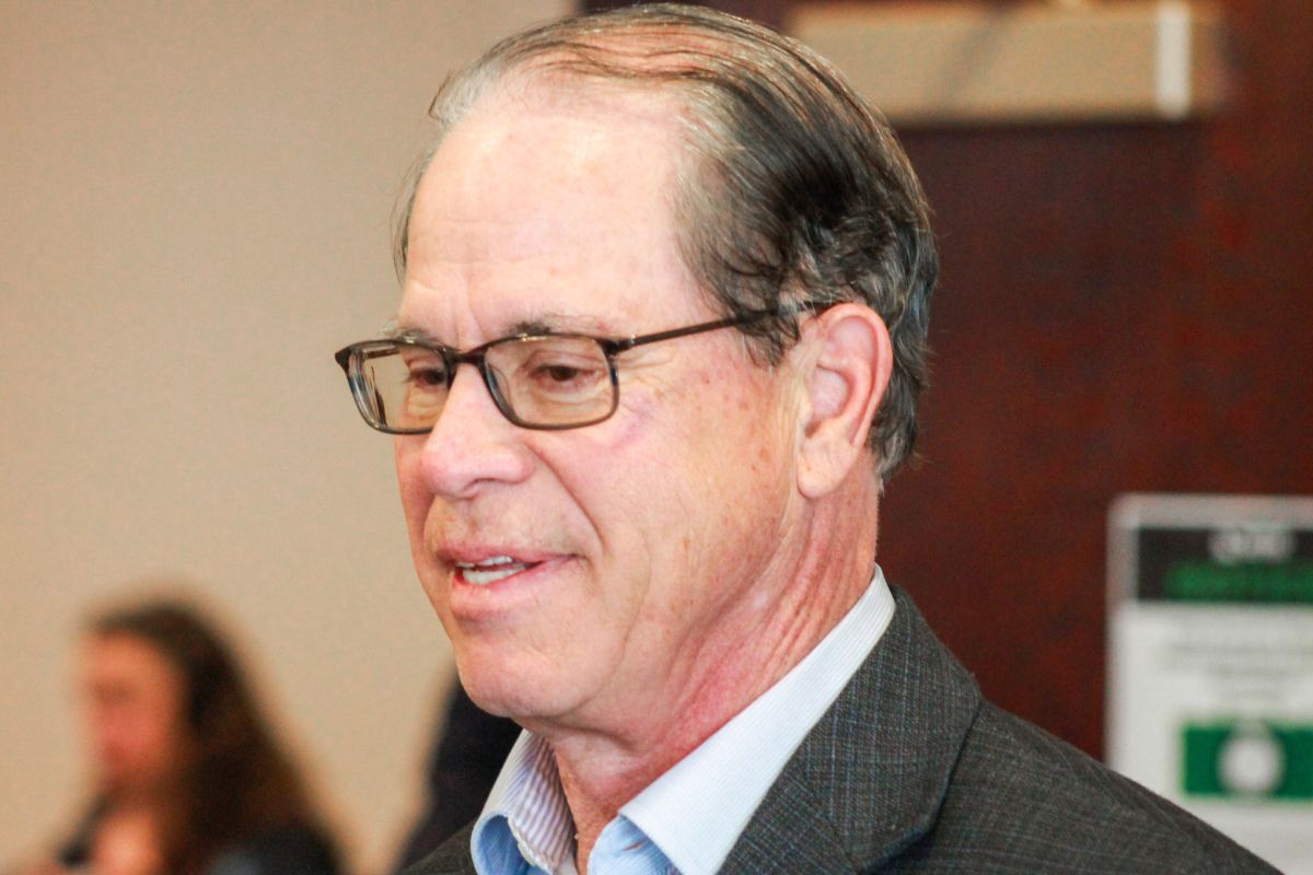 Mike Braun smiles as he speaks to people at a candidate forum. Braun is a White man, balding with dark, graying hair. He is wearing glasses and a gray jacket over a light blue button-down shirt.