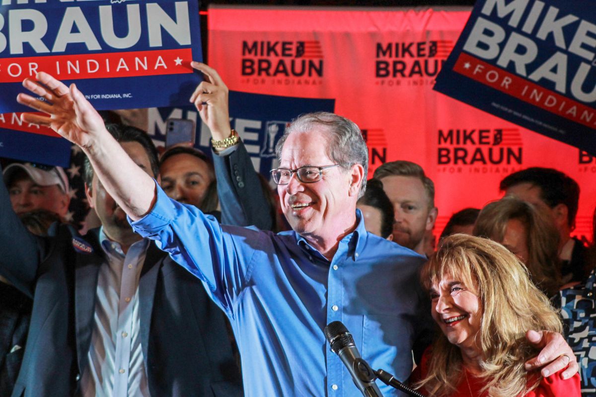 Mike Braun is a White man, balding with gray hair. He is wearing glasses and a blue shirt. Maureen is a White woman with dark blonde hair. She is wearing a red top.