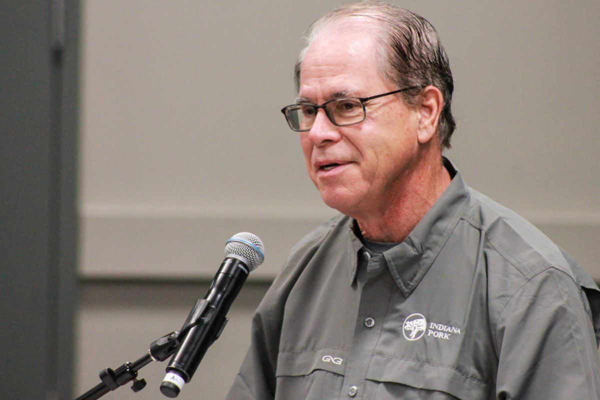 Mike Braun speaks into a microphone. Braun is a White man, balding with dark gray hair. He is wearing glasses and a button-down shirt with the Indiana Pork Producers logo.