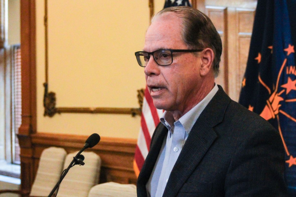 Mike Braun speaks into a microphone in his office. Braun is a White man, balding with gray hair. He is wearing glasses and a dark blazer over a white shirt.