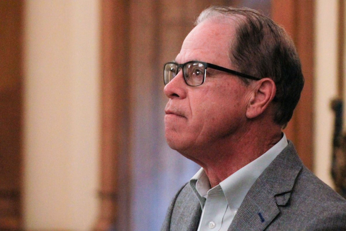 Mike Braun looks upward as he stands in his office. Braun is a White man, balding with gray hair. He is wearing glasses and a gray suit jacket over a white shirt