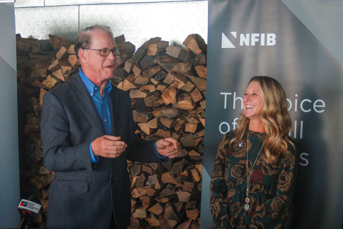 Mike Braun speaks while Natalie Robinson stands beside him. Both are laughing. Braun is a White man with dark, graying hair. He is wearing glasses and a gray jacket over a blue shirt. Robinson is a White woman with blonde hair.