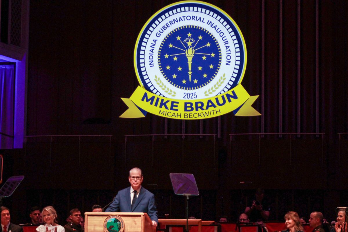 Mike Braun stands on stage at a lectern while a logo for the inauguration ceremony hangs above him. Braun is a White man, balding with gray hair. He is wearing glasses and a suit.