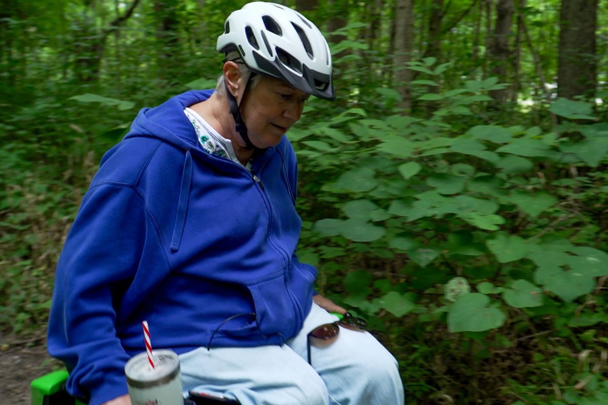 Nancy navigates her trail chair named Peggy in Eagle Creek. She wears a blue zip-up hoodie and light blue pants. She also wears a helmet on the trail with a drink sitting in a cupholder on the chair. Behind her is lush green foliage.