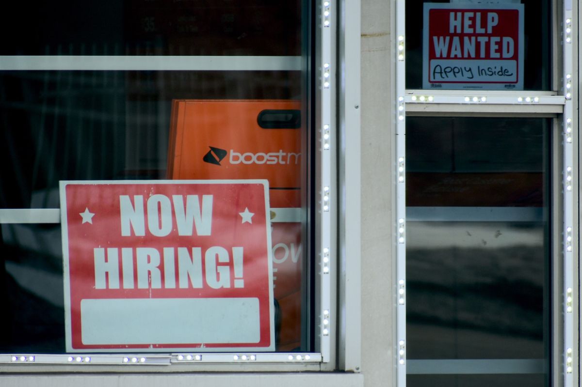 A red and white NOW HIRING sign is placed in the window of a business. To its right, a smaller HELP WANTED sign is wedged into a tall narrow window with Apply Inside written in neat handwriting.