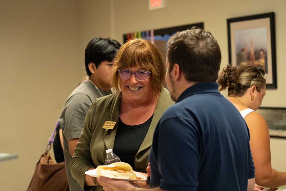 Veronica Pejril wears a dark sage blazer with a name tag, speaking to someone at an event from the Indiana Stonewall Democrats.