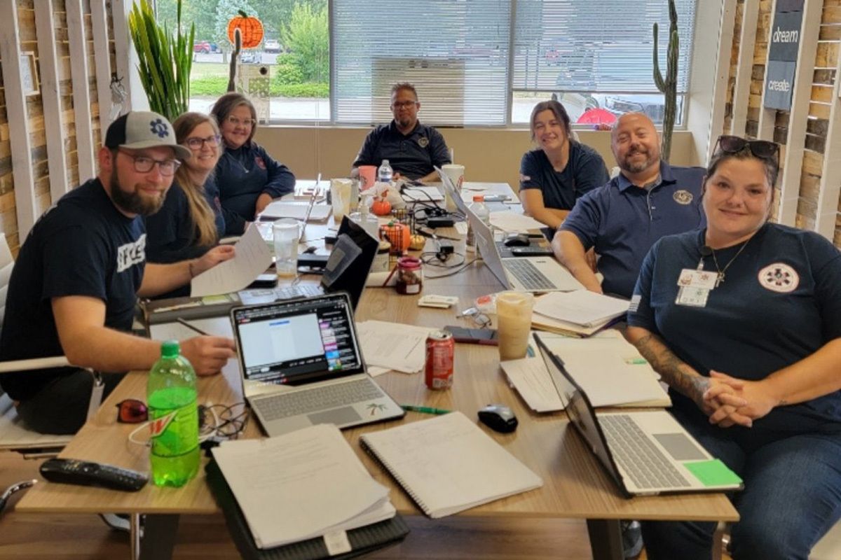 A group of people from Phoenix Paramedic Solutions sit around a table, posing for a camera. The table is covered in papers and laptops.