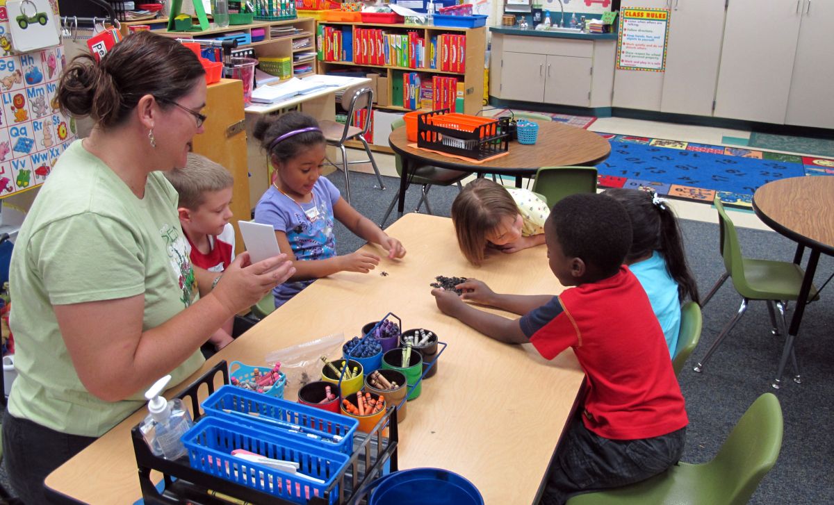 There are five students sitting at a table with there teacher playing a game with each other. 