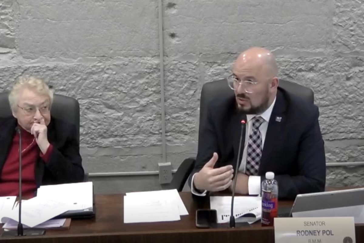 Senator Rodney Pol gestures and speaks into a microphone. Senator Sue Glick watches with her hand against her mouth.