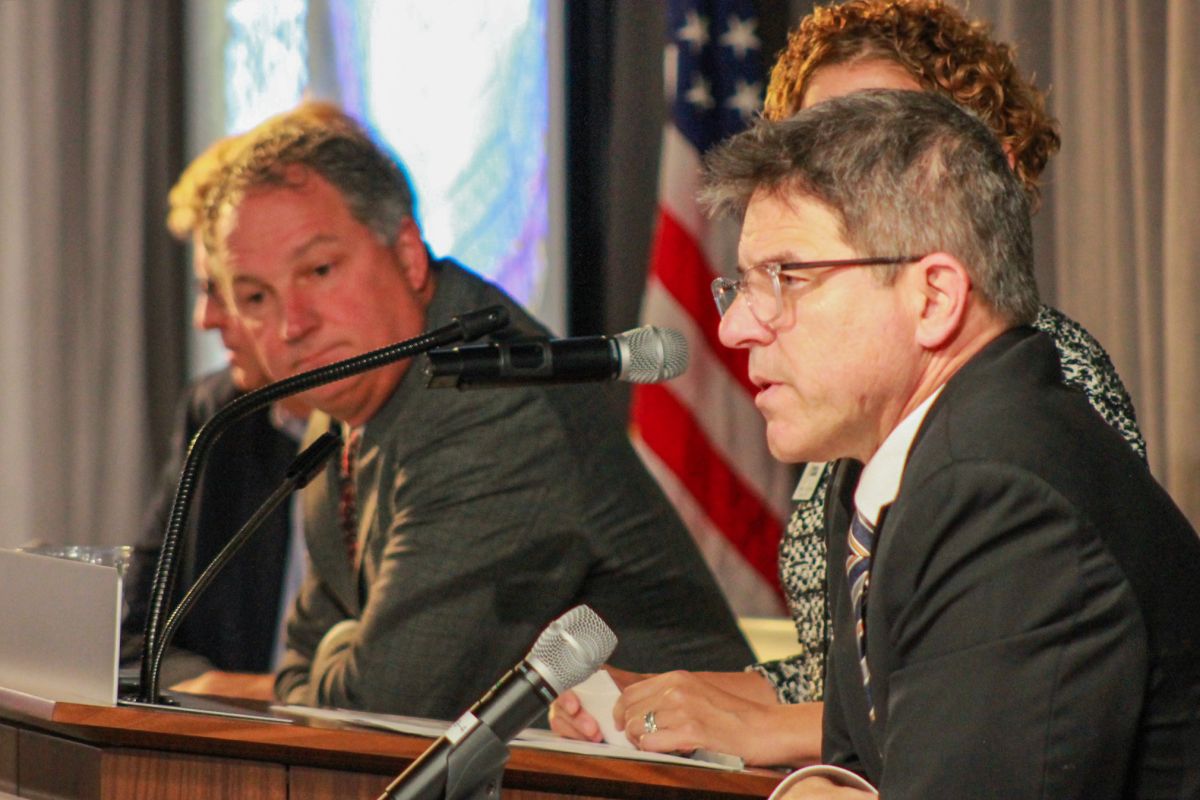 Rodric Bray sits on a stage and talks into a microphone while Todd Huston, also seated, looks on in the background. 