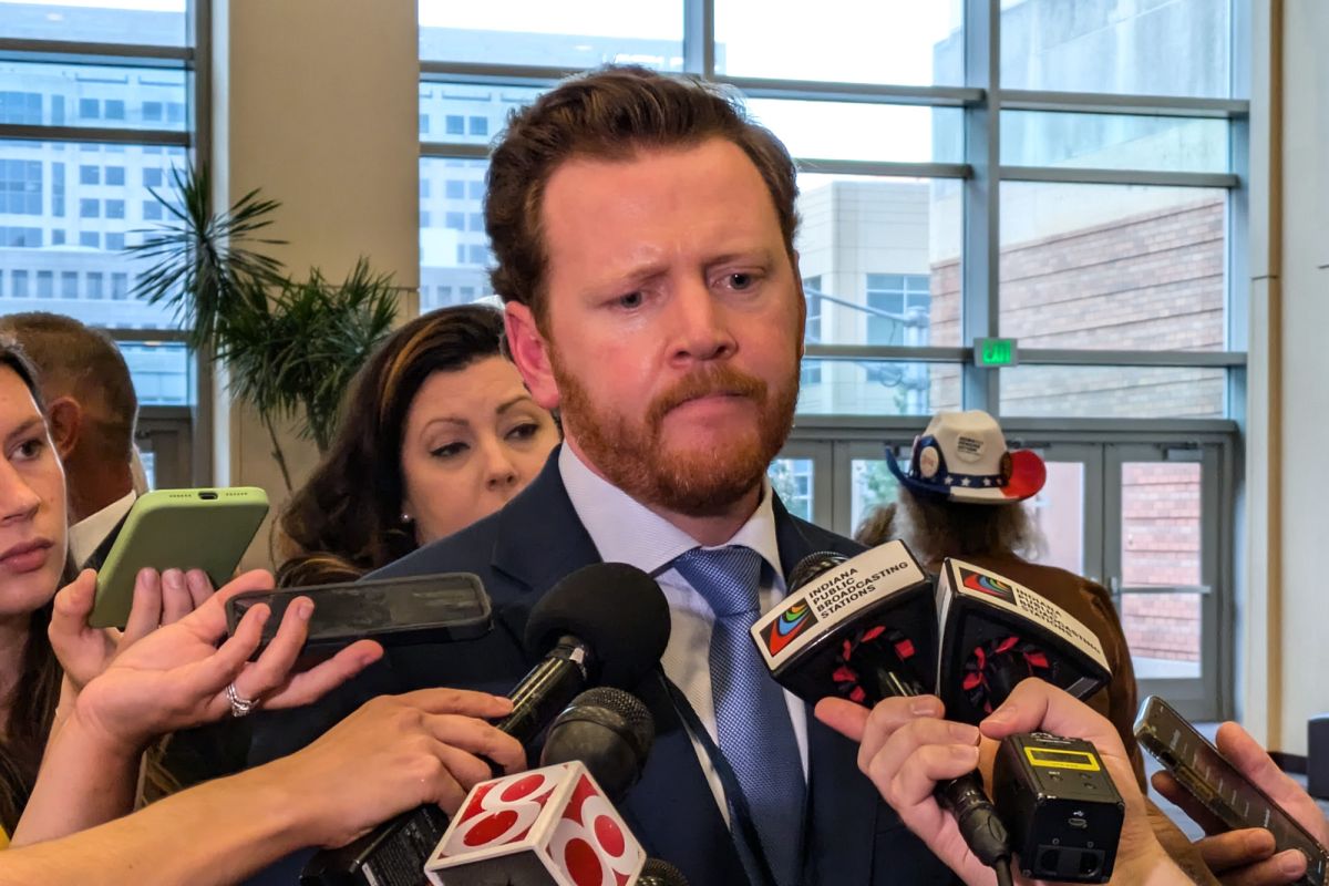 Mike Schmuhl stands in the middle of a press gaggle with phones and microphones surrounding his face to capture his remarks. Schmuhl is a White man with red hair and a beard.